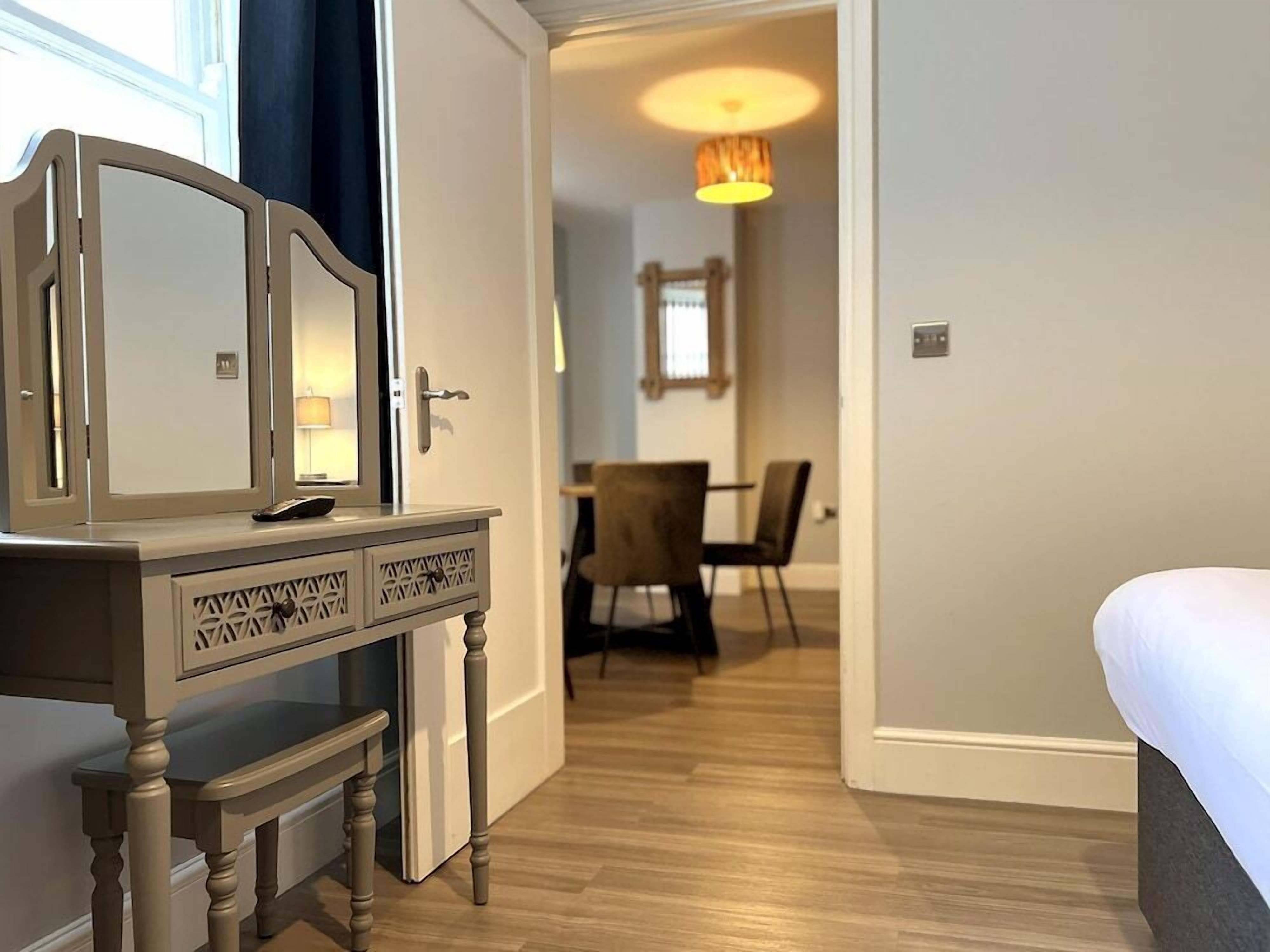 A hotel room with a wooden dresser, a bed, a dining area with chairs, and a lamp.