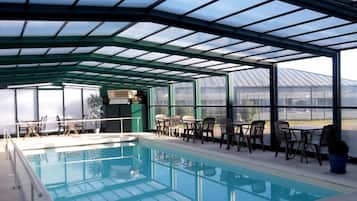 A covered swimming pool with seating areas and a view of a greenhouse in the background.