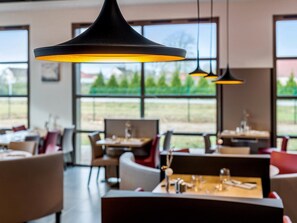 Un interior de restaurante moderno con ventanas amplias, lámparas colgantes negras y mobiliario contemporáneo.