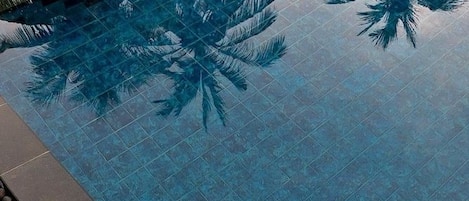 An infinity pool with palm tree reflections, surrounded by dark tiles and a pebble border.