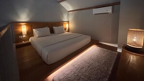 A bedroom with a sloped ceiling, wooden headboard, a bed with white linens, a bedside table with a lamp, and a small rug. - Umadewi Surf and Suites (Pekutatan)