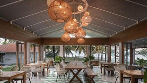 A spacious indoor area with a large wooden table and chairs, surrounded by large windows offering views of palm trees and a building. - Umadewi Surf and Suites (Pekutatan)