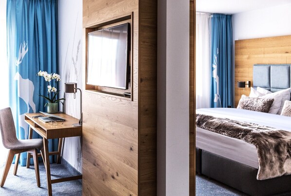 Une chambre d’hôtel avec un bureau en bois, une chaise, un lit avec une tête de lit grise et une grande fenêtre avec des rideaux.