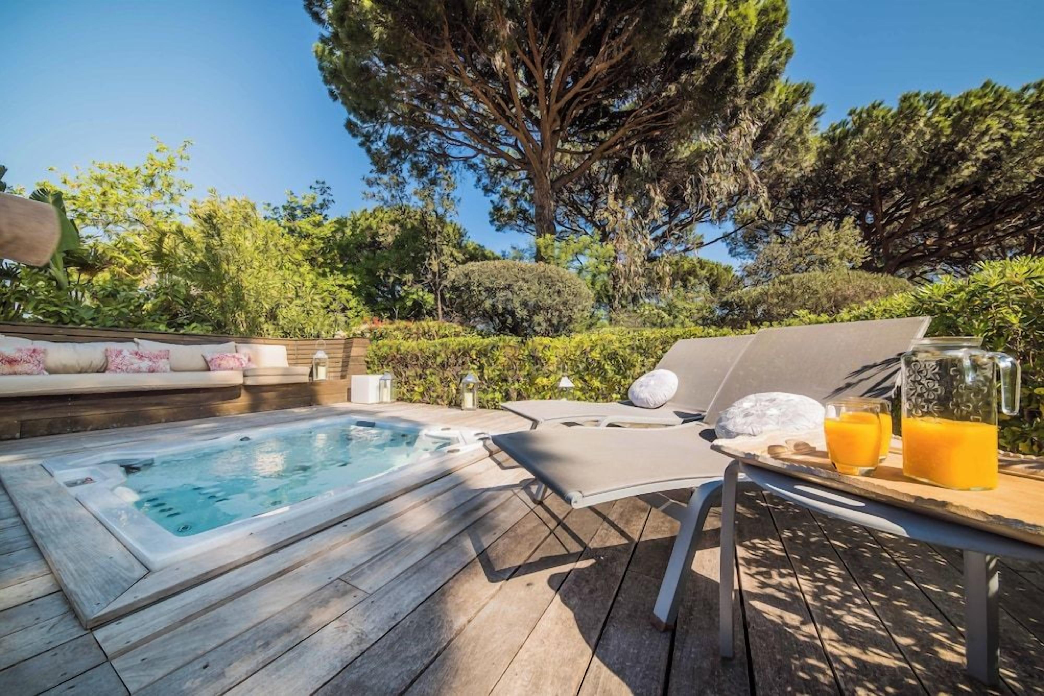 A hot tub on a wooden deck with lounge chairs and a table with drinks.