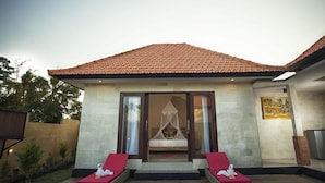 Eine Villa mit Ziegeldach, Glastür-Eingang, einem Himmelbett und zwei roten Liegestühlen draußen.