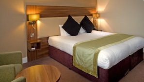 Une chambre d’hôtel avec un grand lit, deux lampes de chevet, une table de chevet en bois, un fauteuil vert et une table ronde en bois.