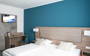 Quarto de hotel com uma cama grande, uma escrivaninha com cadeira, televisão na parede e uma parede de destaque em tom verde-água.