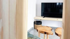 A room with a television on a wooden shelf, a woven basket, and two wooden stools. - Irida Luxury Apartments (Naxos)