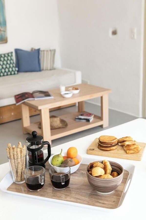 A breakfast tray with coffee, pastries, and fruits on a white table in a modern room with a sofa and a small wooden table in the background. - Irida Luxury Apartments (Naxos)