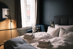 Una habitación de hotel con cama, mesita de noche, lámpara, ventana con cortinas y vistas a edificios.