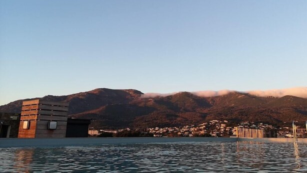A modern building with a pool in the foreground, a mountain range in the background, and a small village across the water. - Hotel and Residence Lido Marana (Furiani)