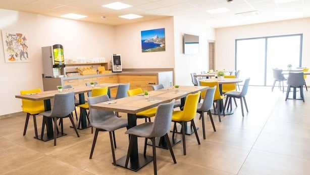Un espace repas moderne avec plusieurs tables et chaises, un coin cuisine équipé d’un réfrigérateur et d’une machine à café, et une grande fenêtre qui laisse entrer la lumière naturelle.