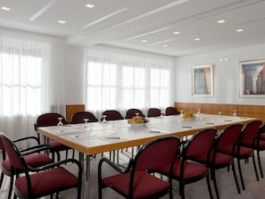 Ein Konferenzraum mit langem Tisch, roten Stühlen, weißen Vorhängen und Deckenleuchten.