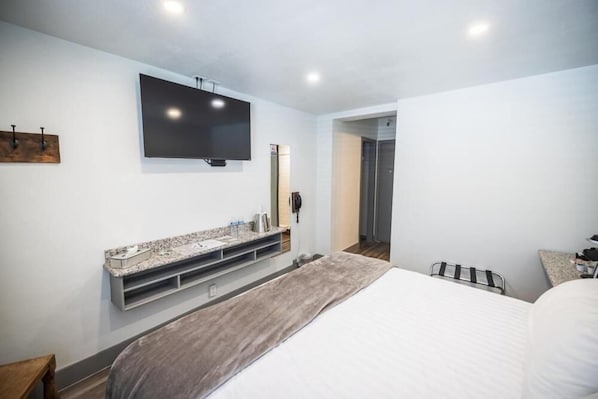 A modern hotel room with a large bed, a flat-screen TV mounted on the wall, a built-in vanity with a mirror, and a small seating area with a bench.