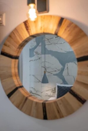 Een ronde spiegel met houten lijst toont een kamer met een marmeren wand en een hangende lamp.