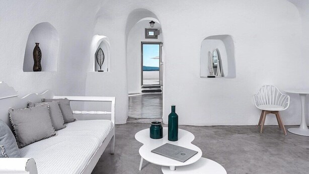 Une pièce moderne et minimaliste, avec un canapé blanc, une petite table blanche et une vue sur la mer, visible à travers une porte en arc de cercle.