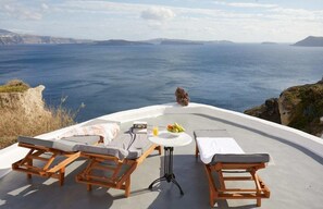 A terrace with wooden lounge chairs and a table, overlooking a scenic ocean view. - Diamond Luxury Suites (Santorini)