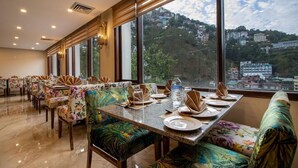 A restaurant with a long table set for dining, large windows offering a view of a hilly landscape, and floral-patterned chairs. - 8fold By Larisa Shimla (Shimla)