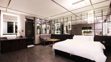 A modern hotel room with a large bed, a vanity with a mirror, a seating area, and a view of the outdoors.