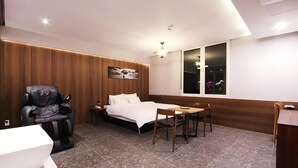 A modern hotel room with a bed, a massage chair, a dining table with chairs, a small desk, and a window with a view of flowers. - Le Idea Hotel (Pohang)