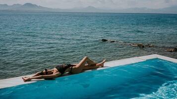A person lying on a poolside lounge chair with a view of the sea and distant mountains.