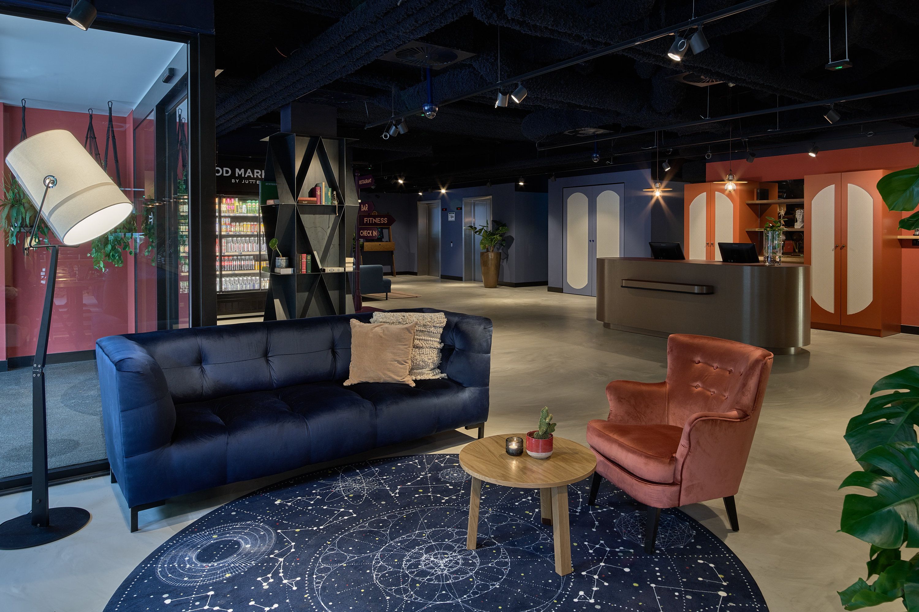 Ein stilvoller Lounge-Bereich mit einem blauen Sofa, einem rosa Sessel und einem Couchtisch. Ein Bücherregal mit Büchern und einer Topfpflanze ist ebenfalls vorhanden.