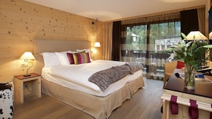 Een hotelkamer met een houten hoofdbord, een bed met meerdere kussens, een nachtkastje met een lamp, een bureau met een computer, en uitzicht op groen buiten het raam.
