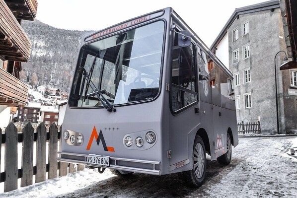 Un autobus est garé dans un village enneigé, avec des bâtiments en bois et une montagne en arrière-plan.