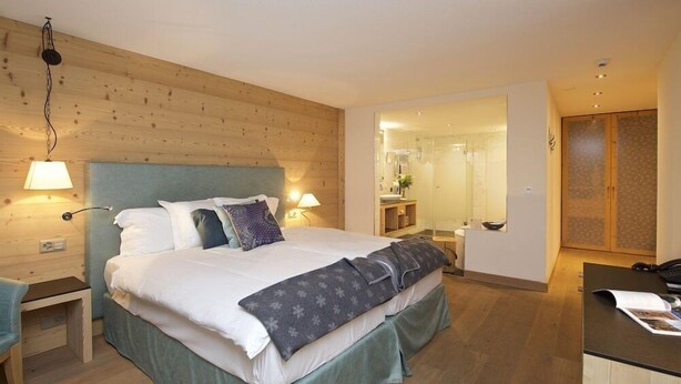 Een hotelkamer met een houten hoofdeinde, een bed met witte lakens, twee bedlampen, een bureau met telefoon, en een badkamer zichtbaar door een geopende deur.