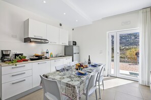 A modern kitchen with white cabinets, a dining table set with plates, glasses, and a bottle, and a view of a hillside through a glass door. - Villa Al Fiume - La Terrazza Dei Sogni in Torbole sul Garda, Torbole sul Garda, Italy (Torbole sul Garda)
