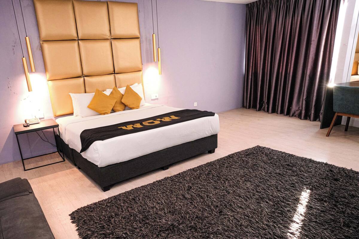 A modern bedroom with a tufted headboard, a large bed with a black and white bedspread, two bedside lamps, a dark shag rug, and a view of a green chair through a window with dark curtains.