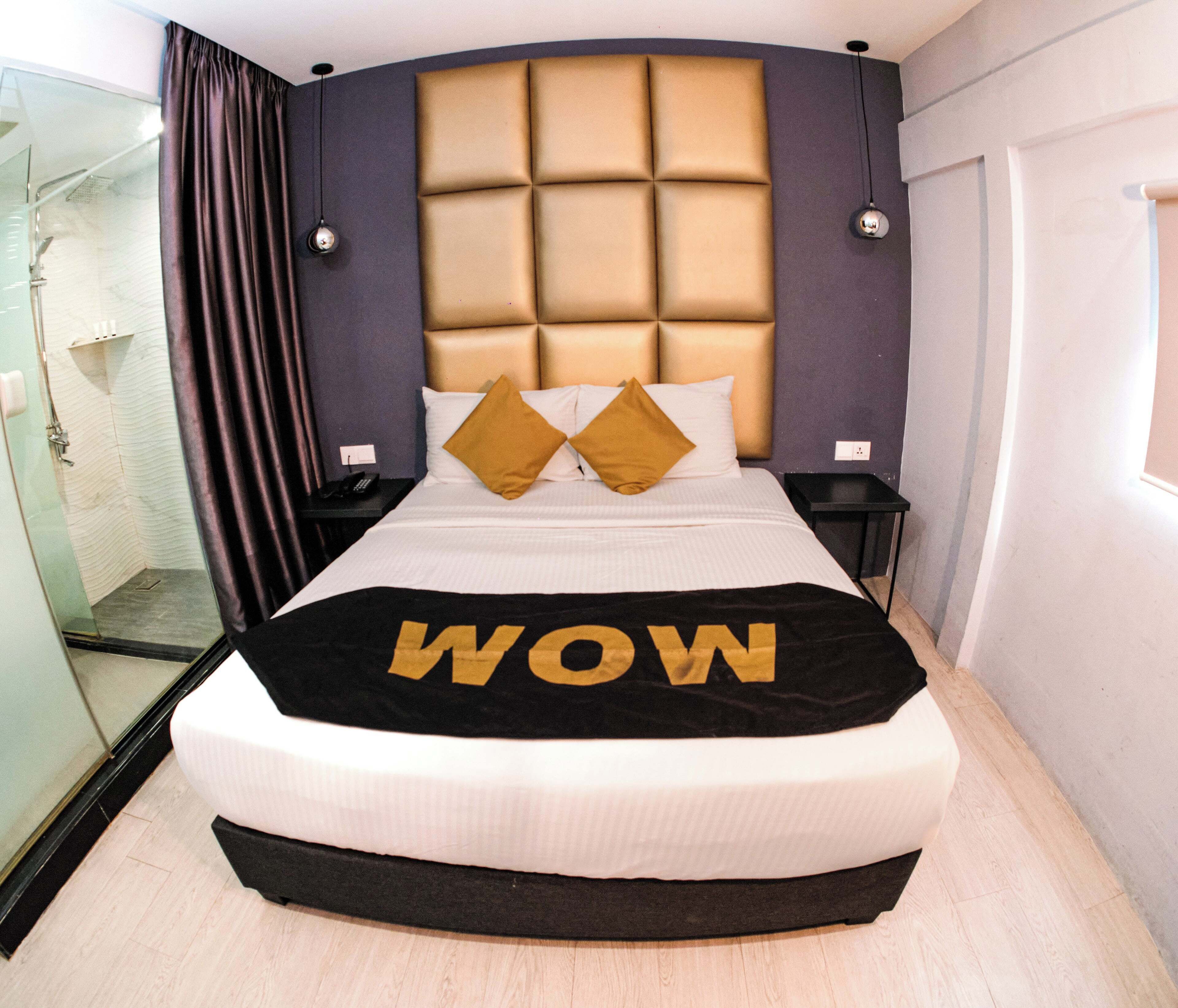 A hotel room with a large bed, a headboard with a padded design, two bedside tables, a glass shower enclosure, and a window.