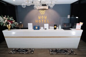 A hotel reception desk with a 'WOW HOTEL' sign, a welcome mat, and a reception counter with various informational brochures and a small plant. - Palette Georgetown Formerly WoW Hotel (George Town)