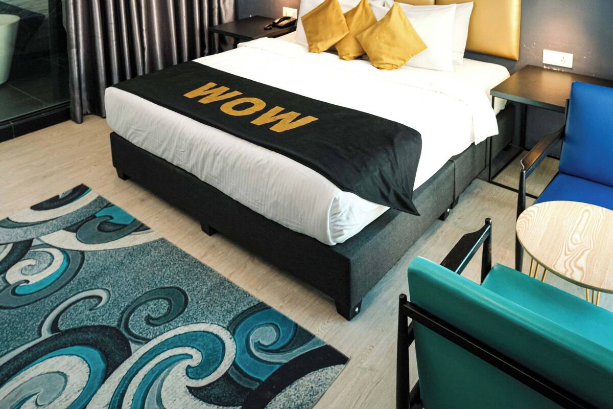 A modern hotel room with a large bed, a headboard, a bedside table, a chair, a small table, and a large window with curtains.