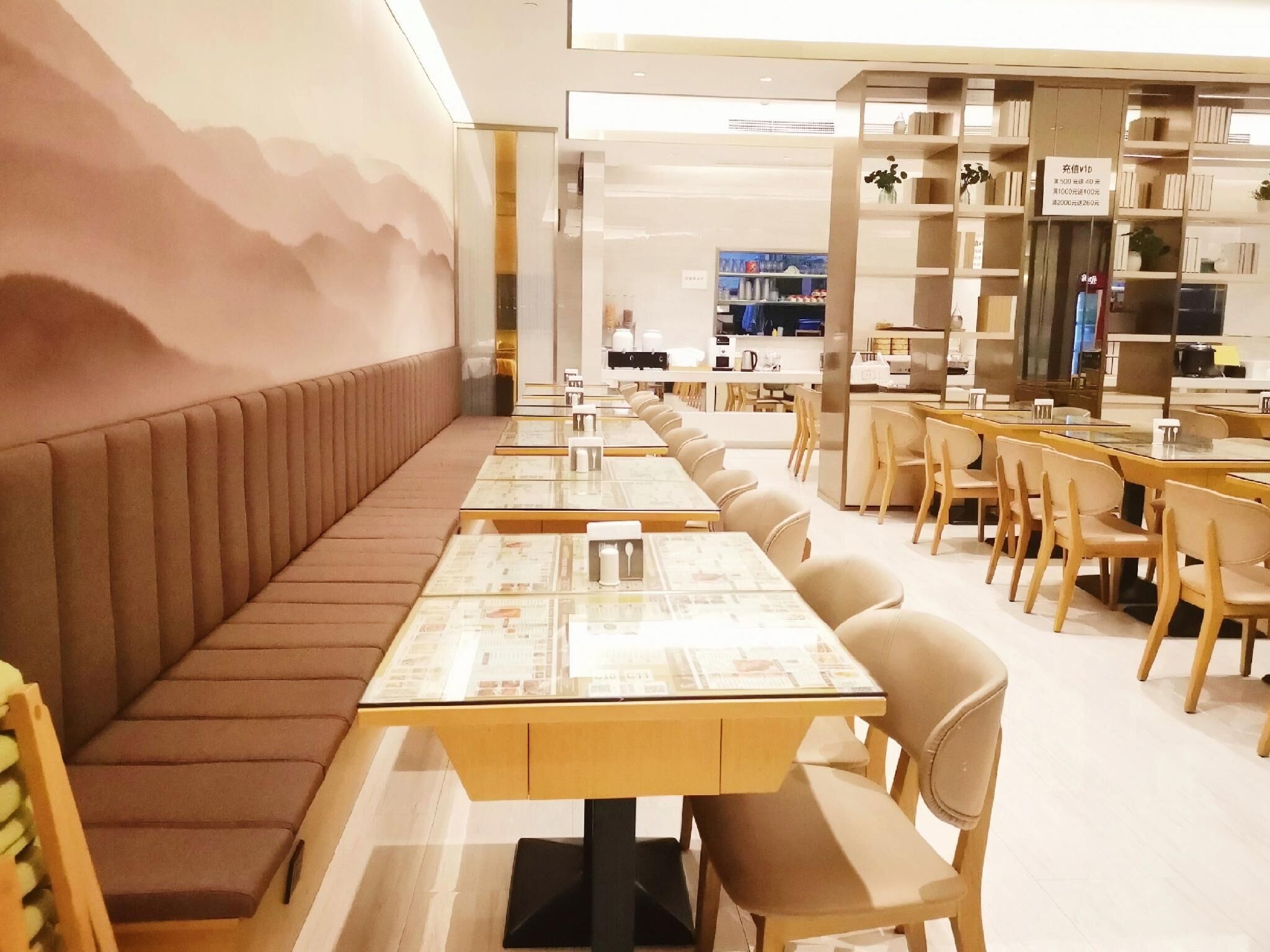 A restaurant with long communal tables and cushioned benches, a kitchen area in the background, and shelves with books and decorative items.