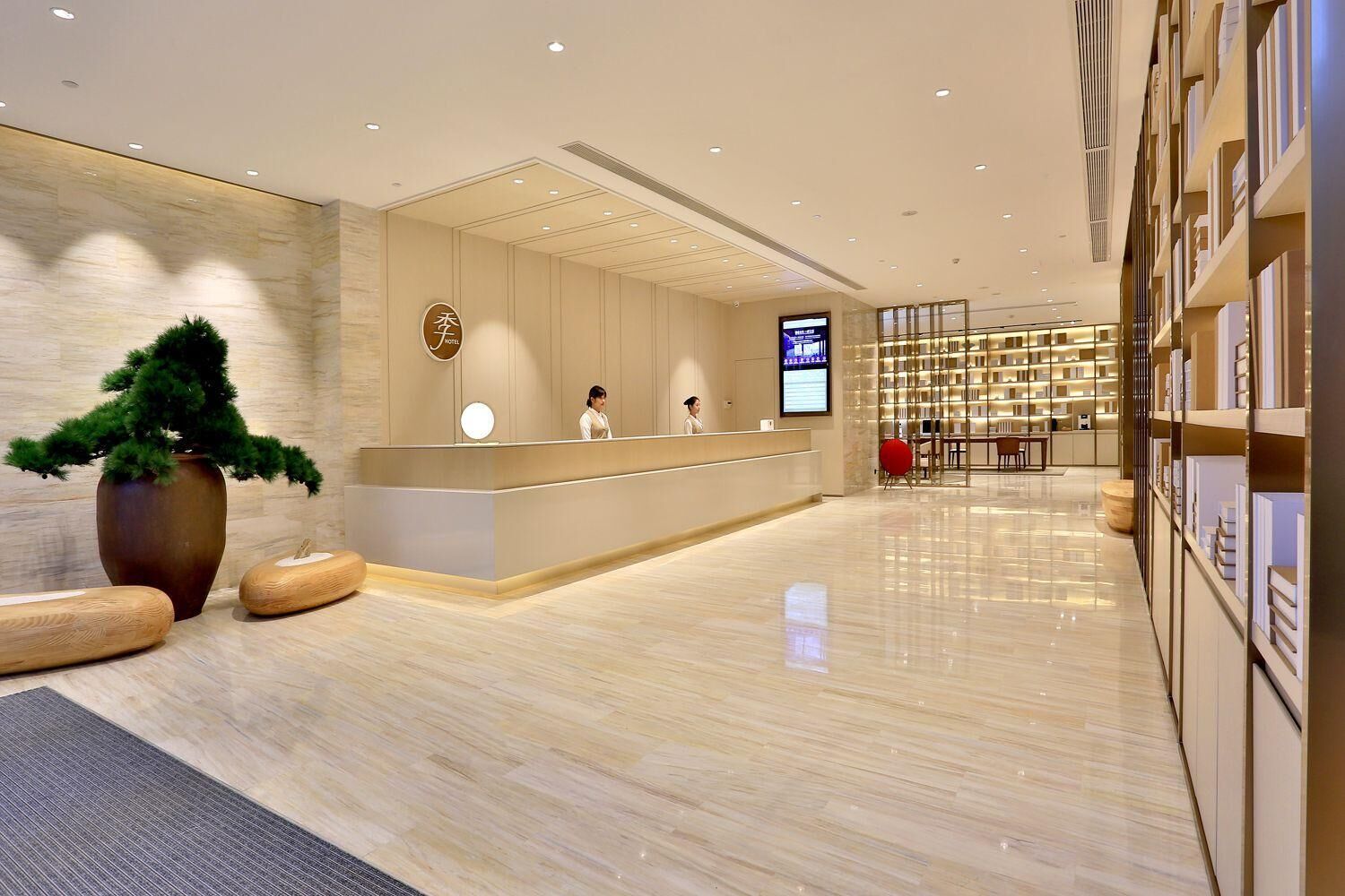 A modern hotel reception area with a reception desk, a large potted plant, and a digital screen displaying information.