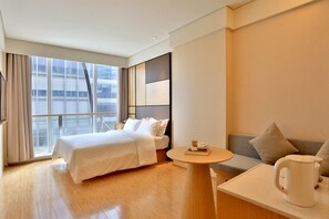 Habitación de hotel con cama, sofá, una mesita con hervidor y vistas a la ciudad a través de amplios ventanales.
