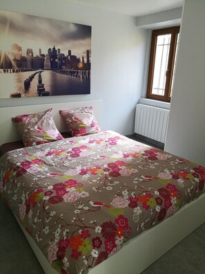 A bedroom with a bed, floral bedding, a cityscape painting, and a window with a view. - Le Cocon des Monts dOr (Saint-Germain-au-Mont-d'Or)