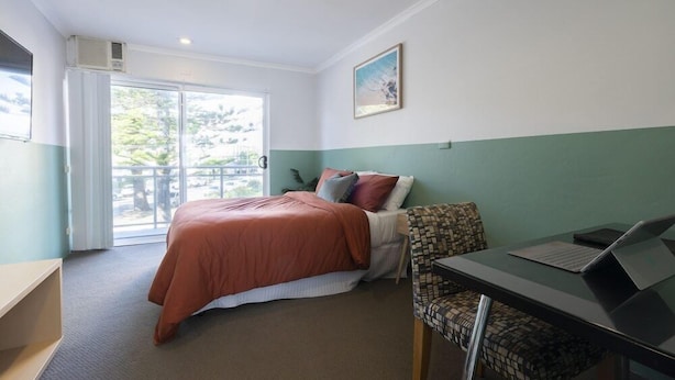 A hotel room with a bed, a desk with a laptop, a chair, and a window with a view of greenery. - Manly Designer Studios (Sydney)