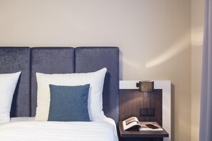 A neatly made bed with white and blue bedding, a wooden nightstand with a lamp, and a book. - Select Hotel Elmshorn (Elmshorn)