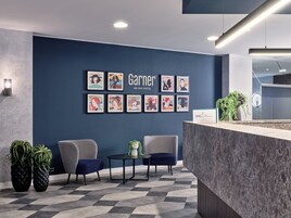 A hotel reception area with a dark blue wall, a reception desk, two chairs, a small table, and framed artwork.