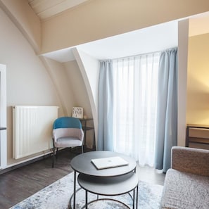 A modern loft bedroom with a sloped ceiling, a round table, a chair, a radiator, and a window with curtains. - Select Hotel Elmshorn (Elmshorn)