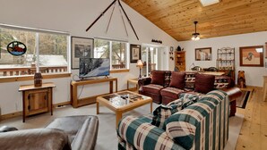 A cozy living room with wooden ceilings, a large window, a fireplace, and a sofa with patterned cushions. - Ceasar Retreat at Big Bear - Cozy Mountain Alpine Cottage (Big Bear City)