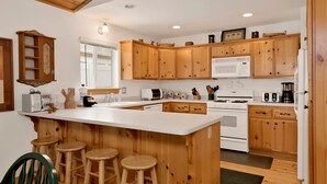 A kitchen with wooden cabinets, a central island with stools, a microwave, a stove, and various kitchen utensils. - Ceasar Retreat at Big Bear - Cozy Mountain Alpine Cottage (Big Bear City)