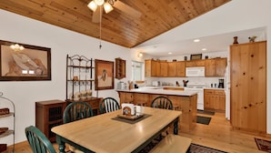 A spacious kitchen with wooden cabinets, a central island, and a dining area with a wooden table and chairs. - Ceasar Retreat at Big Bear - Cozy Mountain Alpine Cottage (Big Bear City)