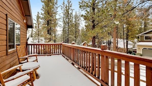 A wooden deck with a railing, snow-covered chairs, and a view of tall pine trees. - Ceasar Retreat at Big Bear - Cozy Mountain Alpine Cottage (Big Bear City)