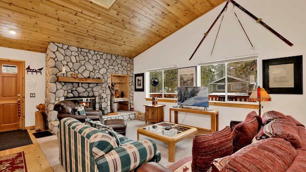 A cozy living room with a stone fireplace, wooden ceiling, and a large window overlooking a snowy landscape. - Ceasar Retreat at Big Bear - Cozy Mountain Alpine Cottage (Big Bear City)