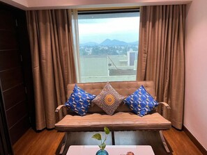 A room with a sofa facing a window with curtains, a small table with a vase and a fruit, and a view of a cityscape. - Fortune Park Vellore (Katpadi)