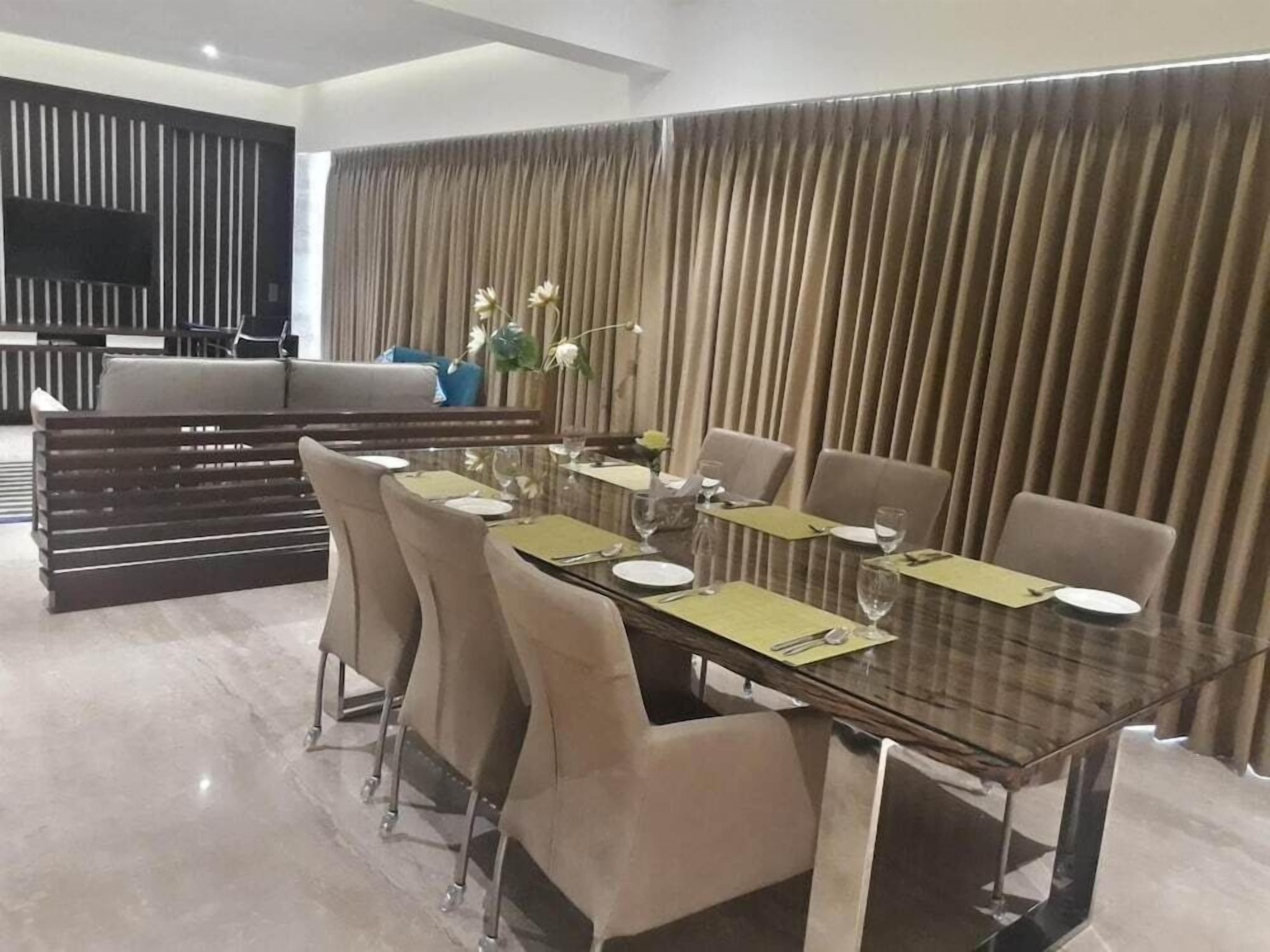 A modern dining area with a glass table, beige chairs, and a backdrop of vertical blinds.