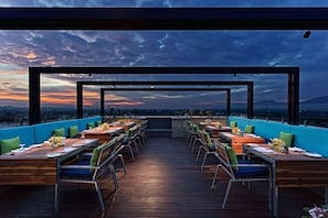 An outdoor dining area with tables and chairs, a wooden deck, and a view of the sunset over a cityscape. - Fortune Park Vellore (Katpadi)